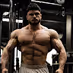 Muscular man posing shirtless in a dimly lit gym with industrial equipment in the background.