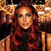 Woman with long red hair and makeup posing against a blurred warm light background.