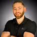 Smiling man with beard and short hair wearing a black polo shirt with arms crossed.
