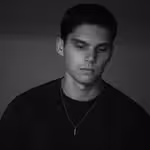 Black and white portrait of a young man with short hair wearing a dark shirt and necklace.