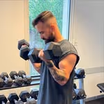 Man lifting dumbbell with one arm in a gym with weights on racks behind him.